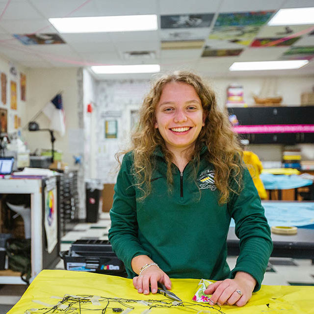 New Life Academy visual arts student working on an art project in the school’s art classroom with colorful student work displayed throughout the room.