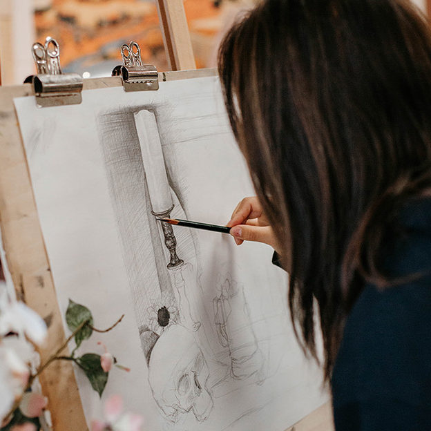 New Life Academy visual arts student sketching a detailed still‑life drawing on an easel in the art classroom.