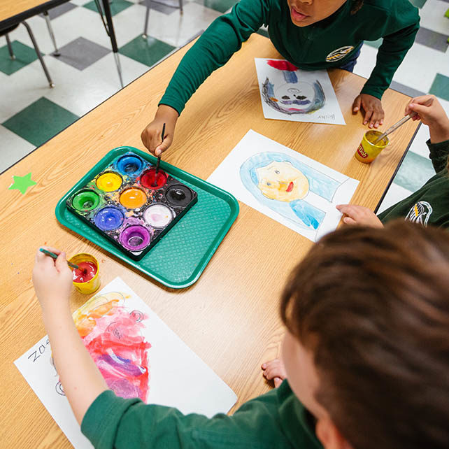New Life Academy lower school students painting with watercolors during art class, using bright colors at a shared classroom table.
