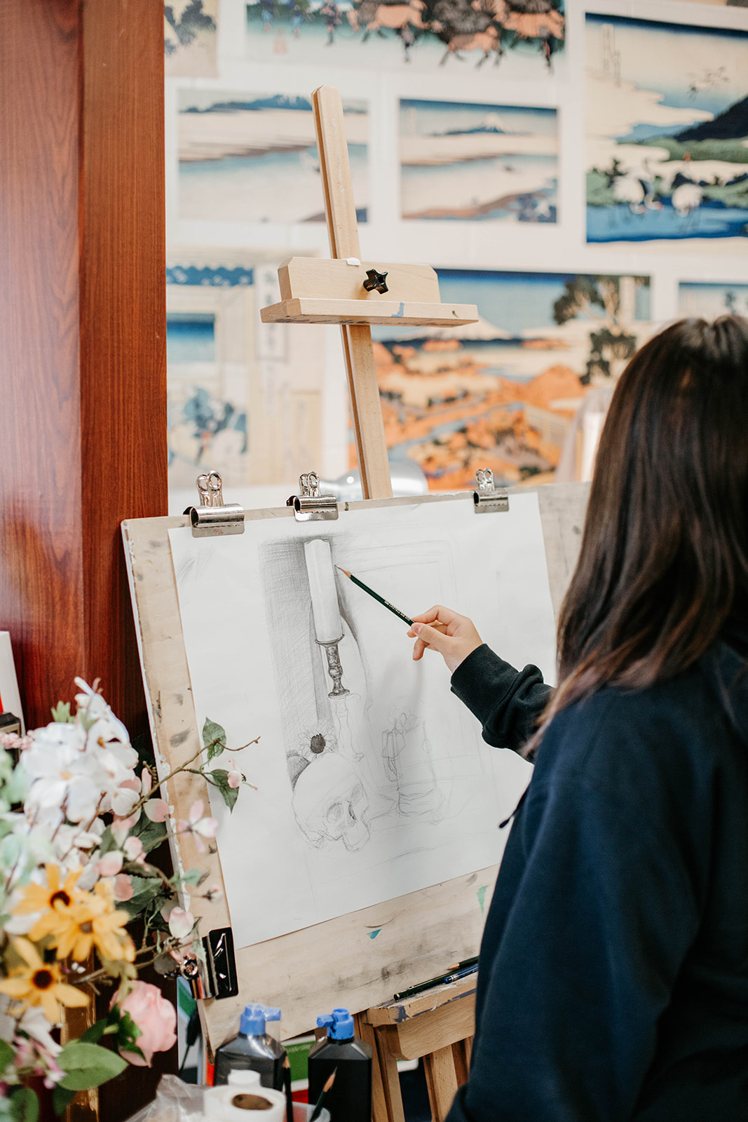 New Life Academy art student sketching at an easel in the visual arts classroom with artwork displayed on the wall.