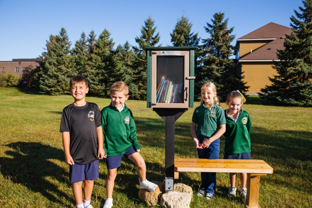 Four New Life Academy students stand smiling beside a Little Free Library filled with books on the school grounds.